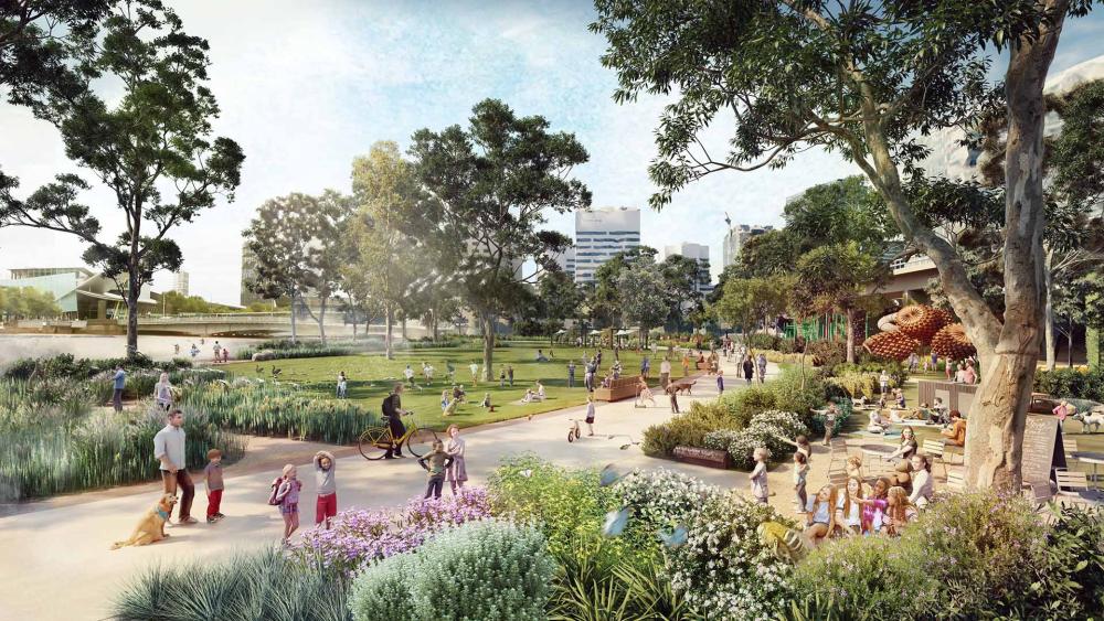 An artist’s impression of what the Batman Park area might become. The image shows a largely green space, with lots of trees and indigenous plants. There is a wide pathway through the centre, being used for families walking the dog, bike riders and people going for a walk. To the left of the pathway, people are sitting on the open grass space closest to the river, while on the right there are more structured seating areas that people are gathered around.  