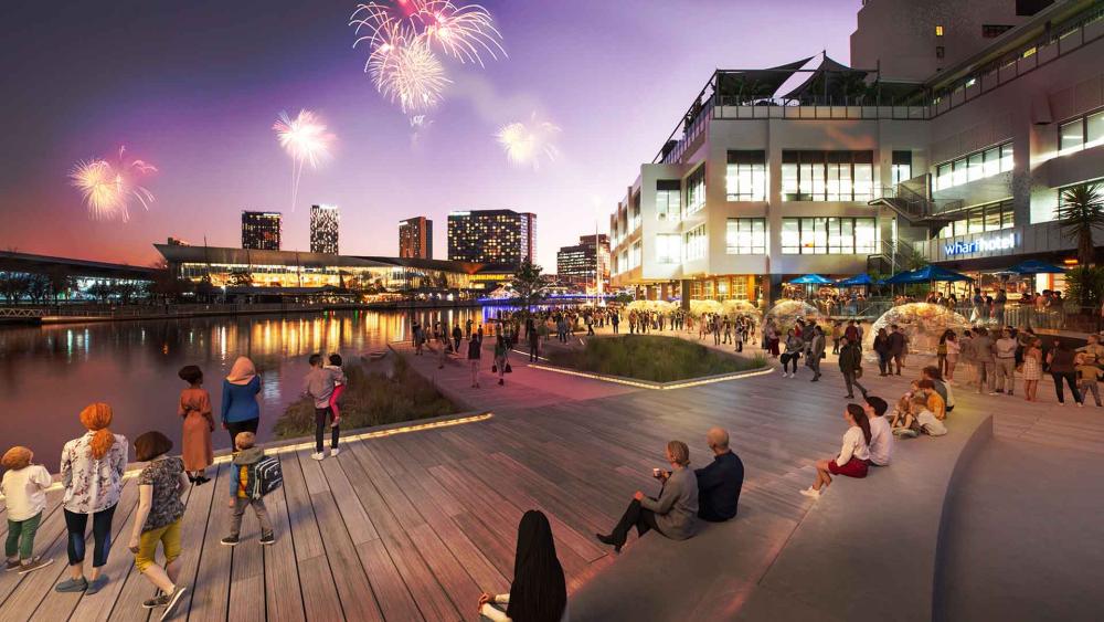 An artist’s impression of what the World Trade Centre Crowne Plaza might become. The image shows onlookers using a wooden boardwalk and steps to observe fireworks that are lighting up the night-time sky over the river. A promenade runs down the right side of the image, busy with pedestrians walking past the hotel-like structure, linking them with the city in the distance.  
